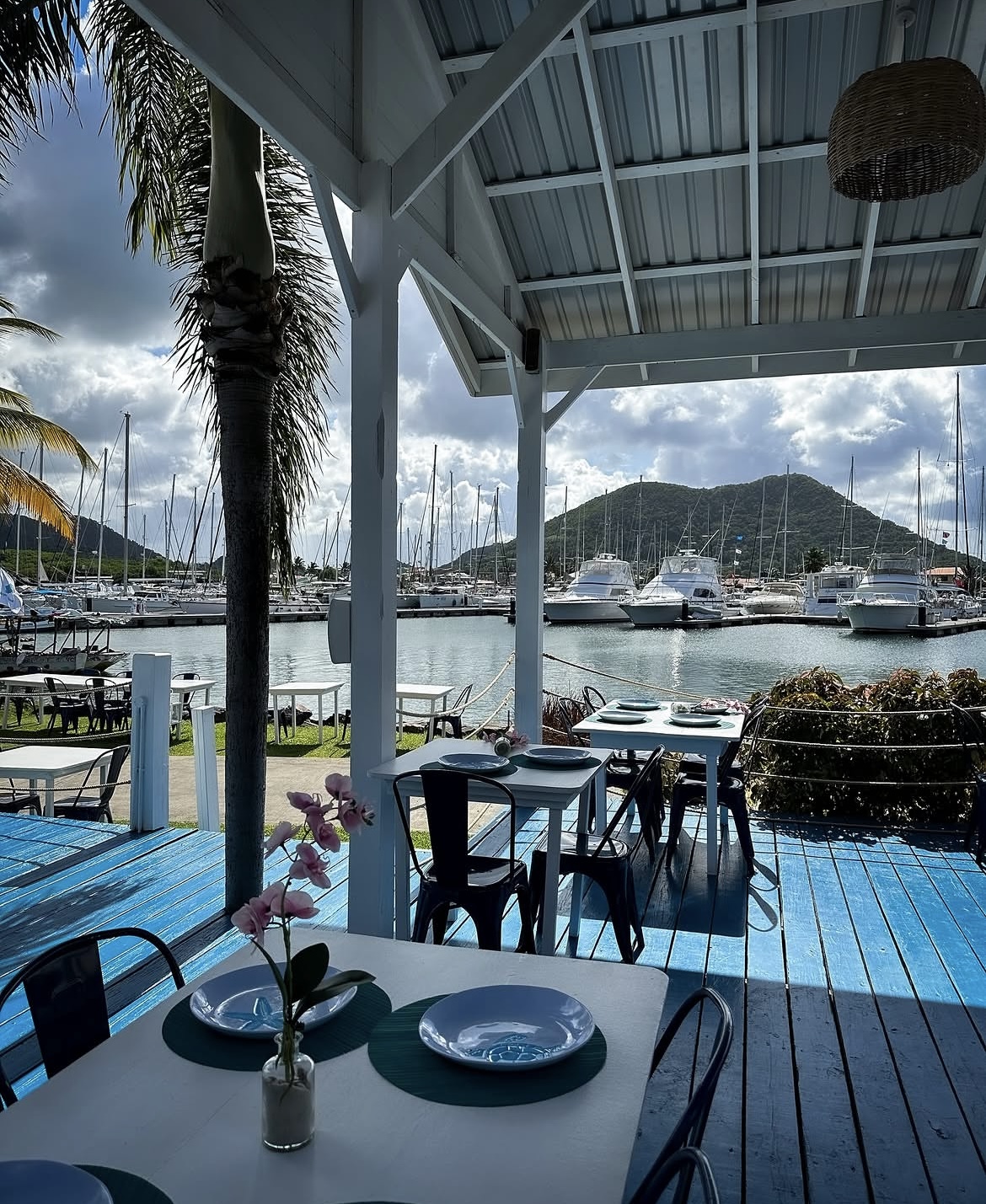 Blue deck tables under the roof with yachts and green hills across the marina